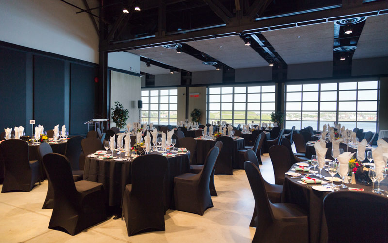 A room with floor to ceiling windows offering a view of the harbour and island and oval banquet tables and chairs set with dining wares.