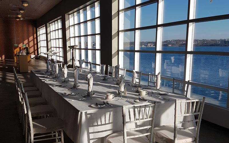 A long table and chairs is set for a banquet for twenty people, with several large windows providing a view of the harbour.