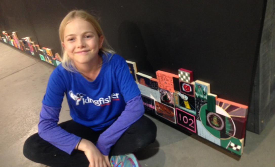 A young girl sits cross-legged in front of a collage of artwork.