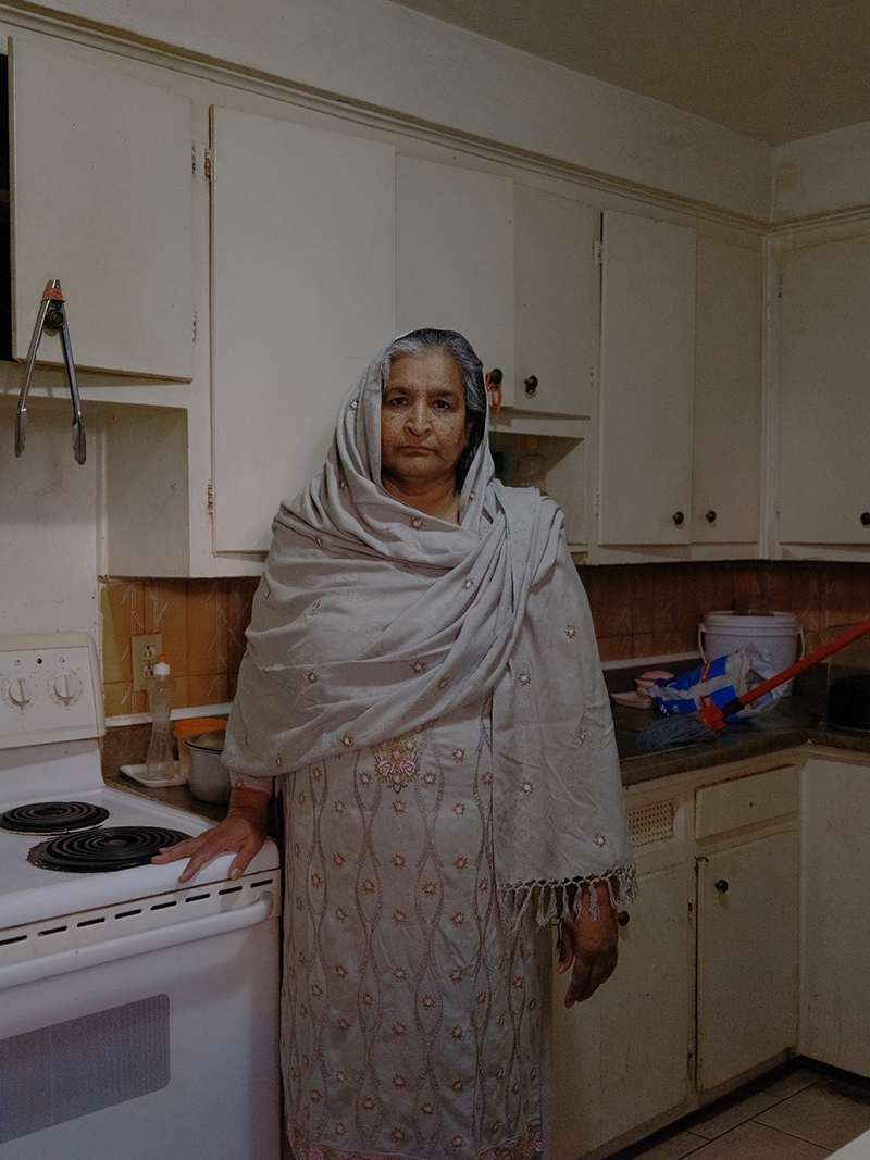 A woman stands in a kitchen, she is a wearing a dress and scarf over her head and has her hand resting on the stove top.