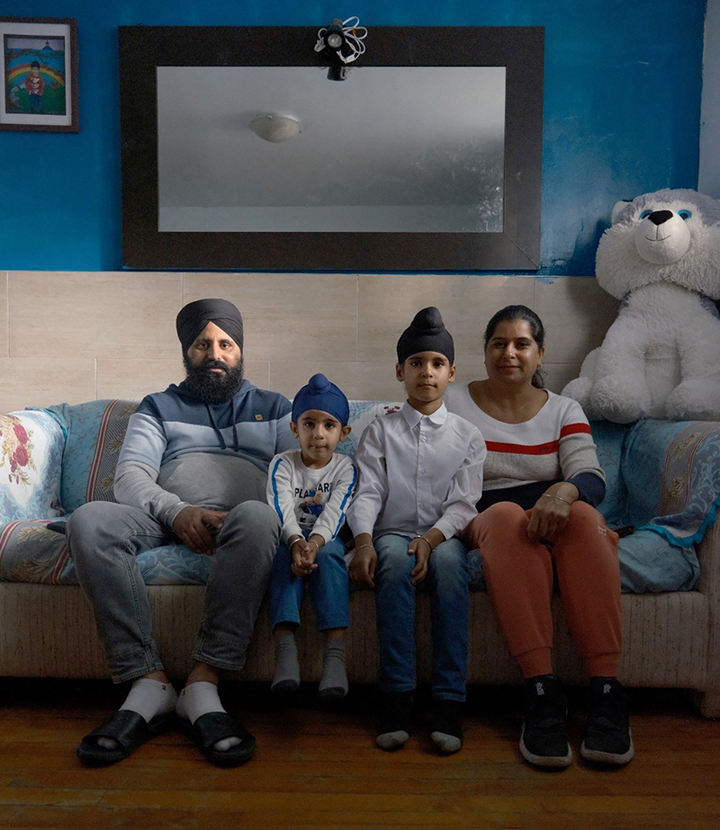 A family of four sit on a couch to have their picture taken; there is a big mirror on the wall behind them.