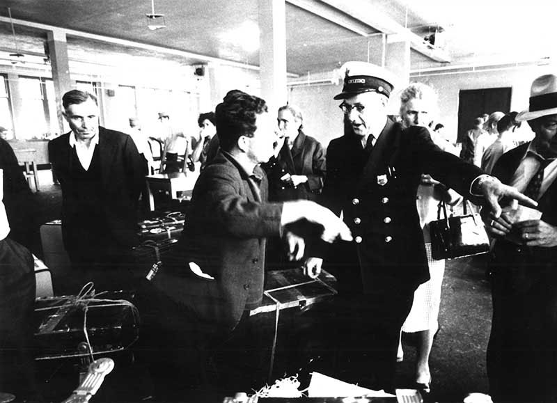 A customs officer is talking to a passenger and pointing to something off-camera.