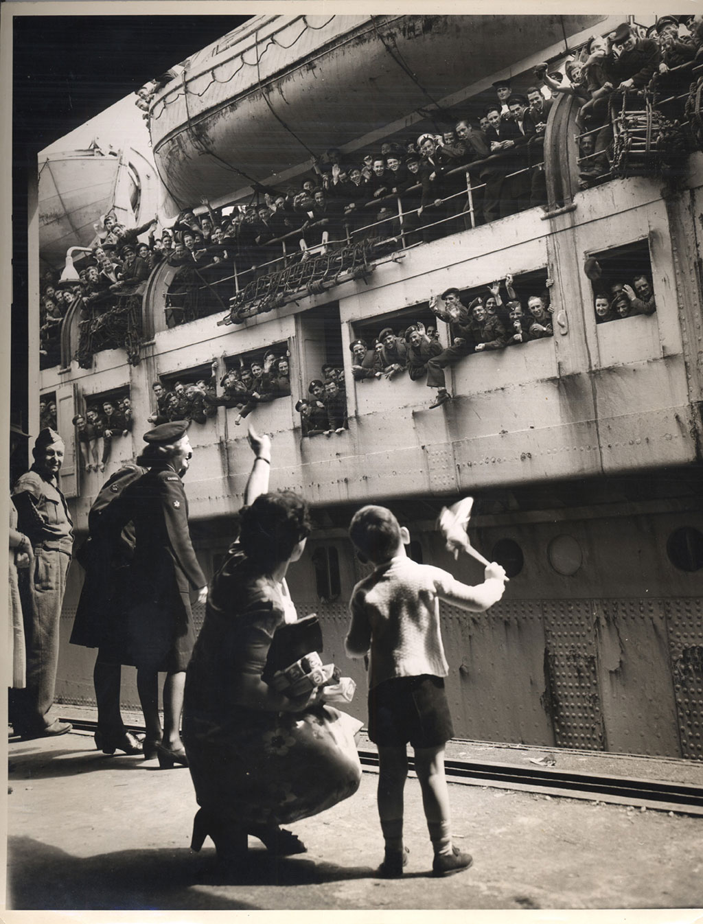Old black and white war-time photo, showing a troop ship docking with hundreds of waving soldiers and a little boy and his mother waving back at them from the platform, while a serviceman looks on.