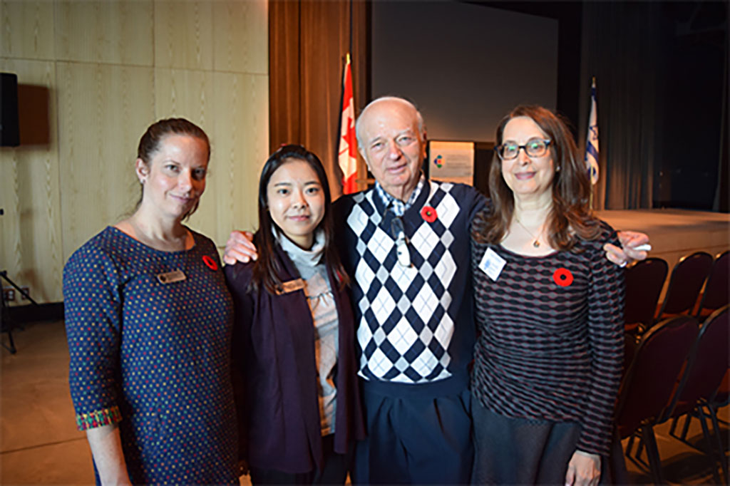 Four people with arms linking stand in a large room, they are all wearing poppies.