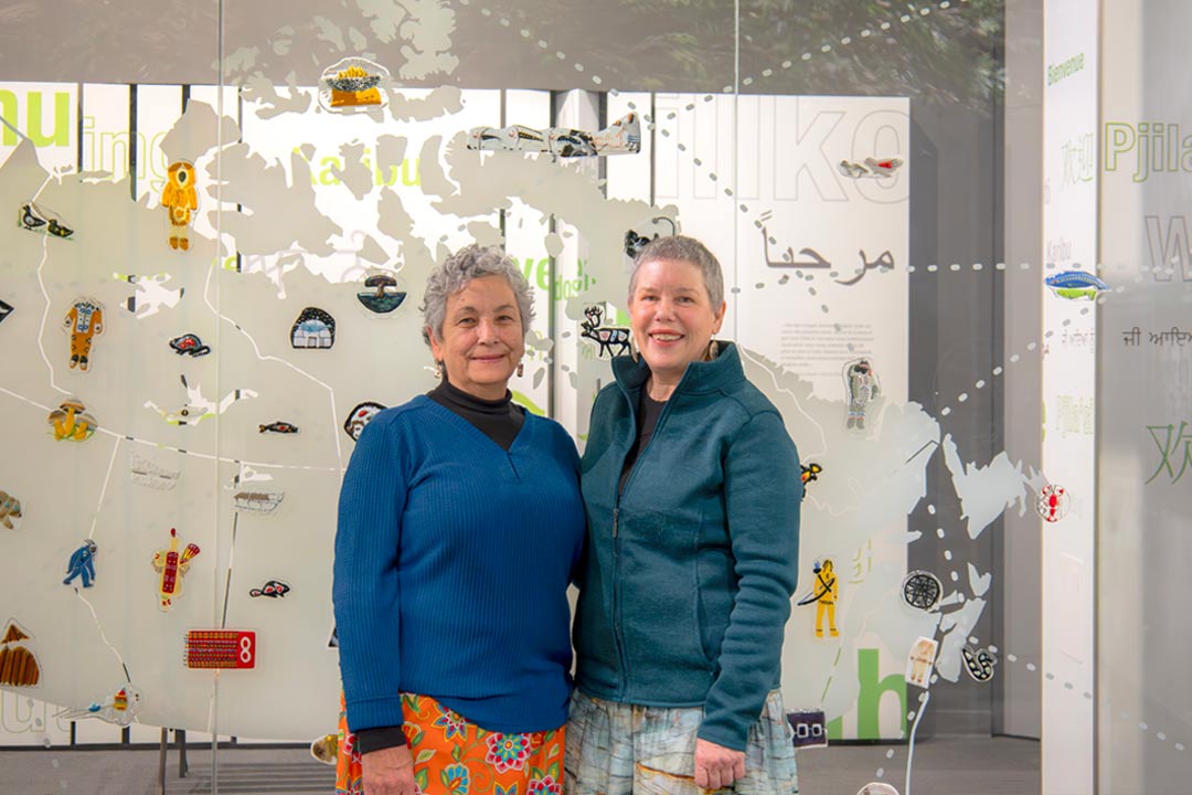 Two smiling Indigenous women stand in front of their glass art installation.