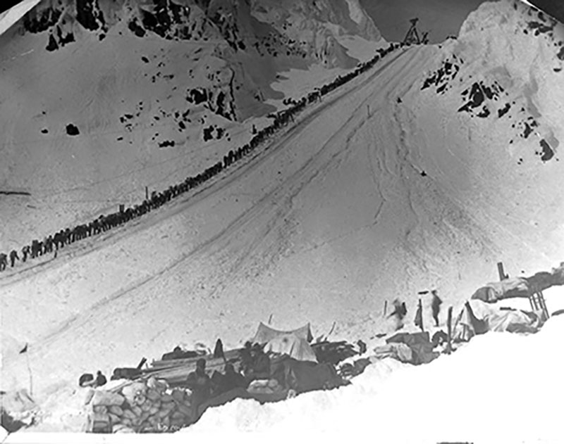 A very long line of people slowly move up a snowy expanse.