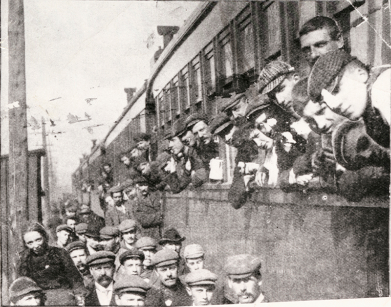 Lots of people milling about on a train platform while others have their heads out the train's windows.