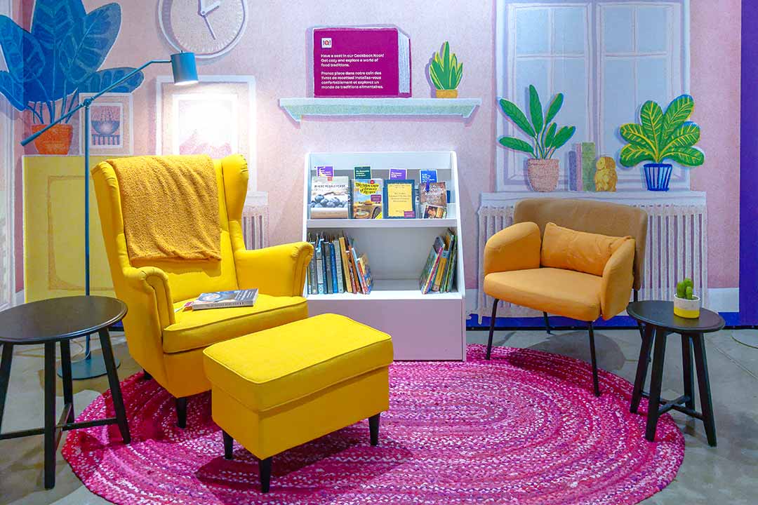 In a mocked-up pink-walled room, a yellow chair and footstool sit on a pink rug beside a bookshelf with two dozen cookbooks.