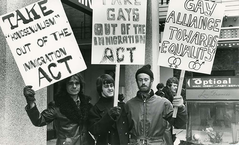 Three men with signs: Take homosexualism out of the Immigration Act, Take gays out of the Immigration Act, and Gay Alliance Towards Equality. They are protesting in front of a commercial building and are dressed for cool weather.