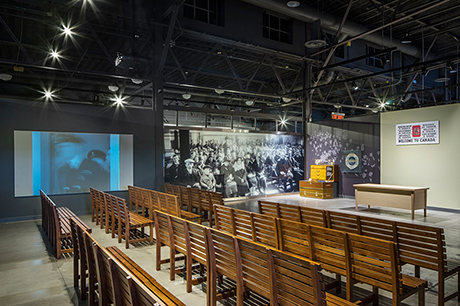 Big hall with wooden benches and large pictures on the wall and a screen showing Pier 21 images.