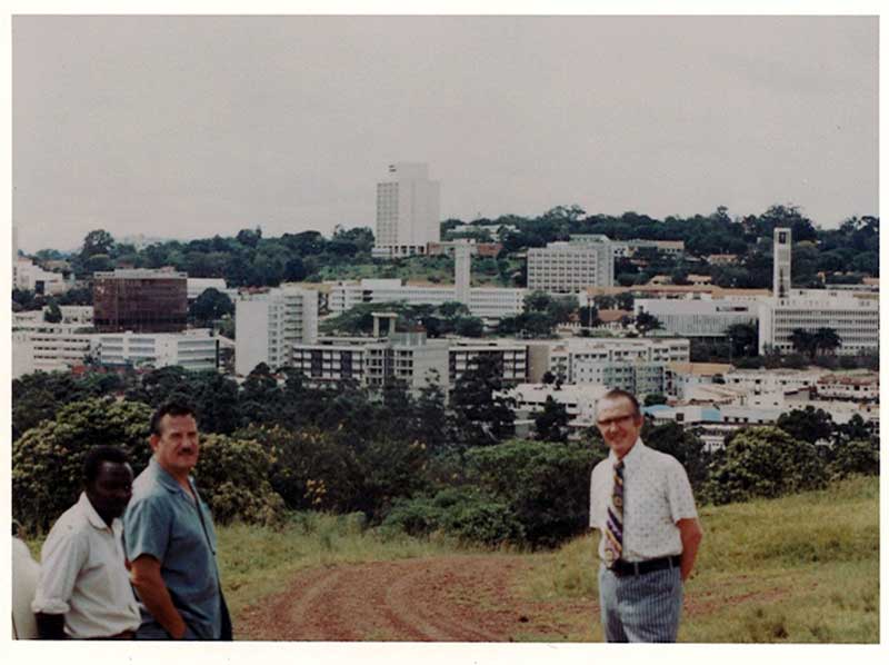 Three men stand on a dirt road with a small city in the background.