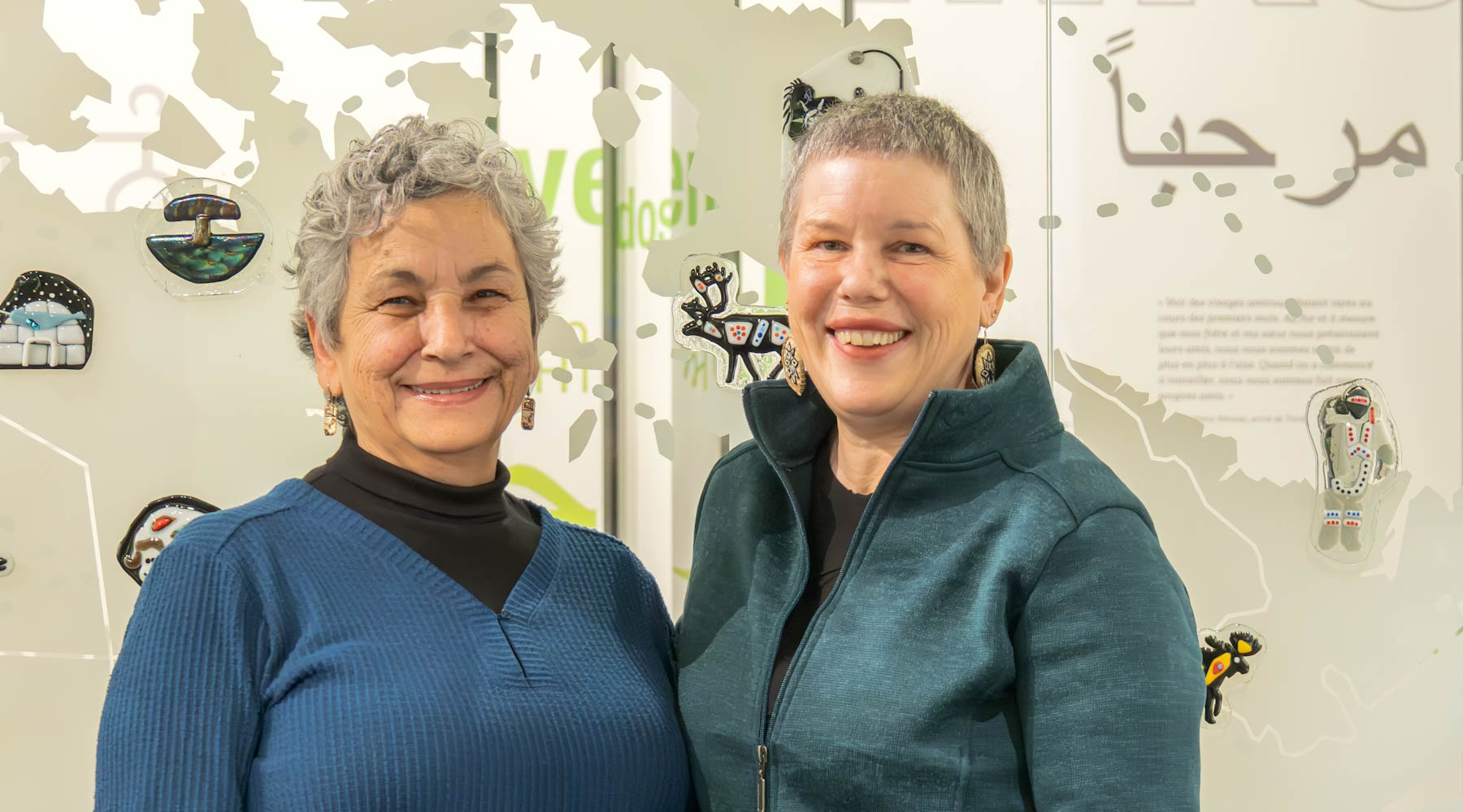 Two Indigenous women with greying hair smile with their arms around each other in front of their artwork.