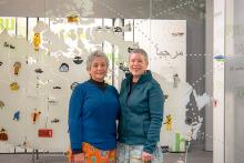 Two smiling Indigenous women stand in front of their glass art installation.