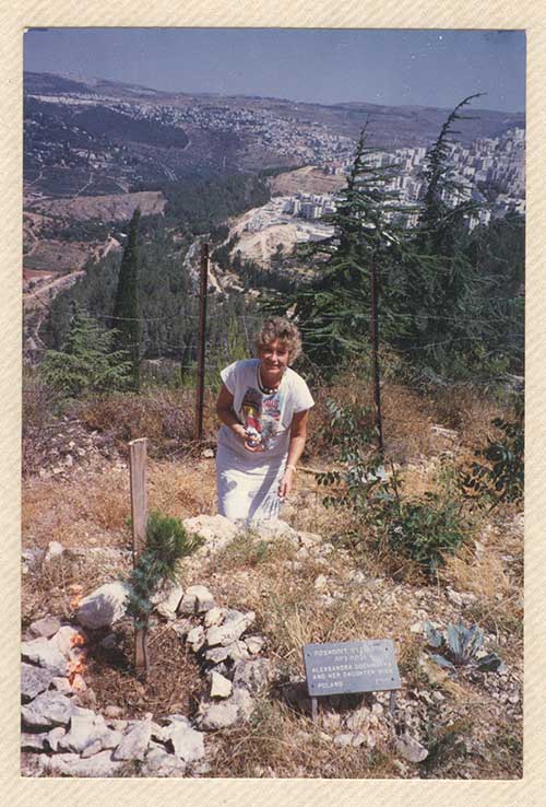 A young woman in a white dress stands on a hill and points at a small tree.