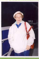 Older Ursula in white jacket, hat and brown purse.