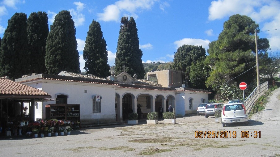 A white cemetery building with flowers in pots and cars parked outside.