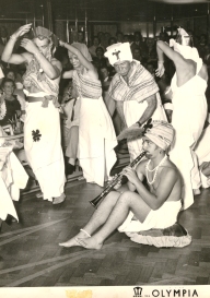 Young man sitting on the floor, playing the flute while costumed people dance around him.