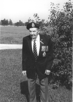 Older Norman, wearing the Legion uniform, sitting on the grass in front of a bush.