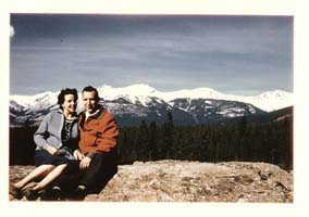 An Italian man and woman sitting on a rocky ledge with trees and snow capped mountains behind them.