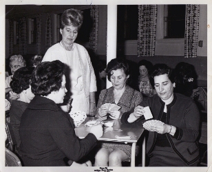 Four women sitting, playing cards, a woman in white standing above.