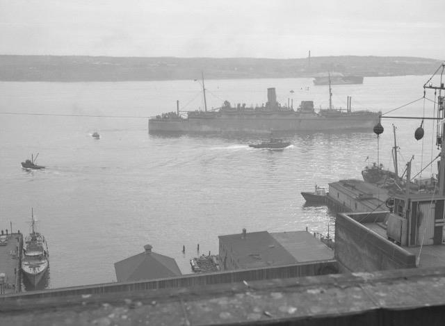 Black and white photo of a large ship in a harbour, with smaller boats around it.