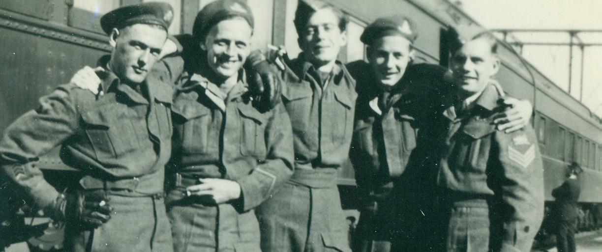Black and white photo of five smiling white men in uniform, with arms around each other and a train in the background.