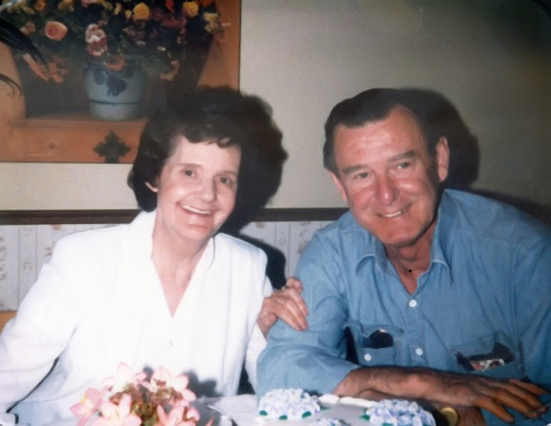 Joan and Les are sitting at a table behind a cake, her arm around his.